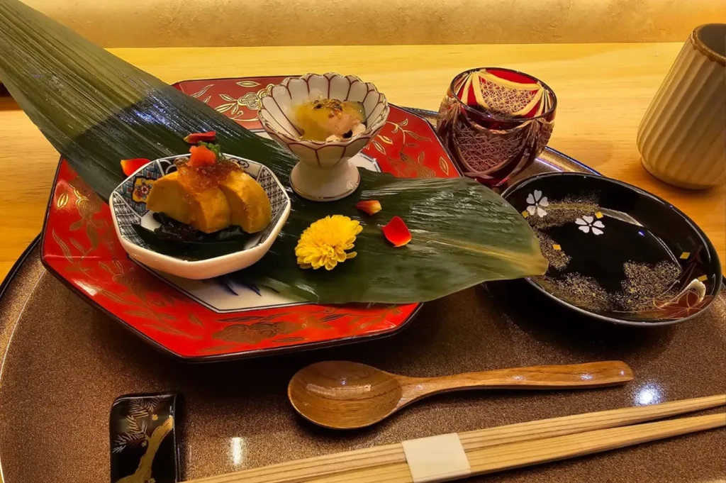 A refined appetizer set is elegantly presented on a large, vibrant red and black lacquer tray, featuring a delicate ceramic bowl with golden-brown glazed pieces topped with red roe, alongside a small, scalloped white bowl containing a glistening, gelatinous dish, all garnished with a bright yellow chrysanthemum and scattered red petals, with a small black side dish holding decorative gold flecks completing the sophisticated composition.