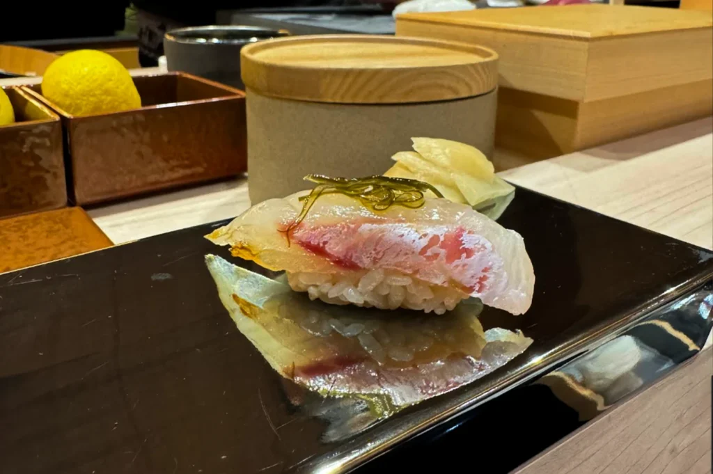 A piece of nigiri sushi topped with a translucent white fish is served on a black rectangular plate. A thin, dark strip of seaweed is placed neatly across the top of the fish, and a small pile of pickled ginger is positioned in the background. In the blurred background, a whole yellow lemon sits in a wooden container next to a rounded wooden box.