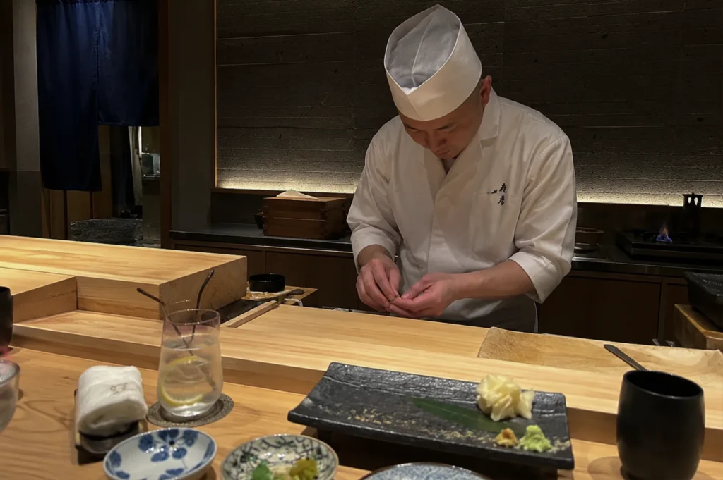 A sushi chef, dressed in a traditional white uniform and hat, is captured in deep concentration as he hand-shapes a piece of sushi behind a long, smooth wooden counter. In the foreground, a dark, rectangular ceramic plate awaits the next serving, accompanied by pickled ginger, wasabi, and a small decorative dish. The setting is a serene, dimly lit dining space with a dark wall behind the chef, highlighting the focus and precision required in preparing high-quality omakase.