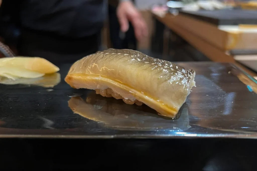 A side-profile close-up highlights a single, glistening piece of anago (sea eel) nigiri sushi resting on a reflective, dark metallic surface. The tender, curved piece of cooked eel features a beautiful gradient from silvery-grey to golden-yellow, lightly brushed with a sweet soy glaze that catches the light over a small bed of seasoned sushi rice. In the soft-focus background, a slice of pickled ginger sits to the left, while the faint silhouette of a chef working at a wooden counter is visible.