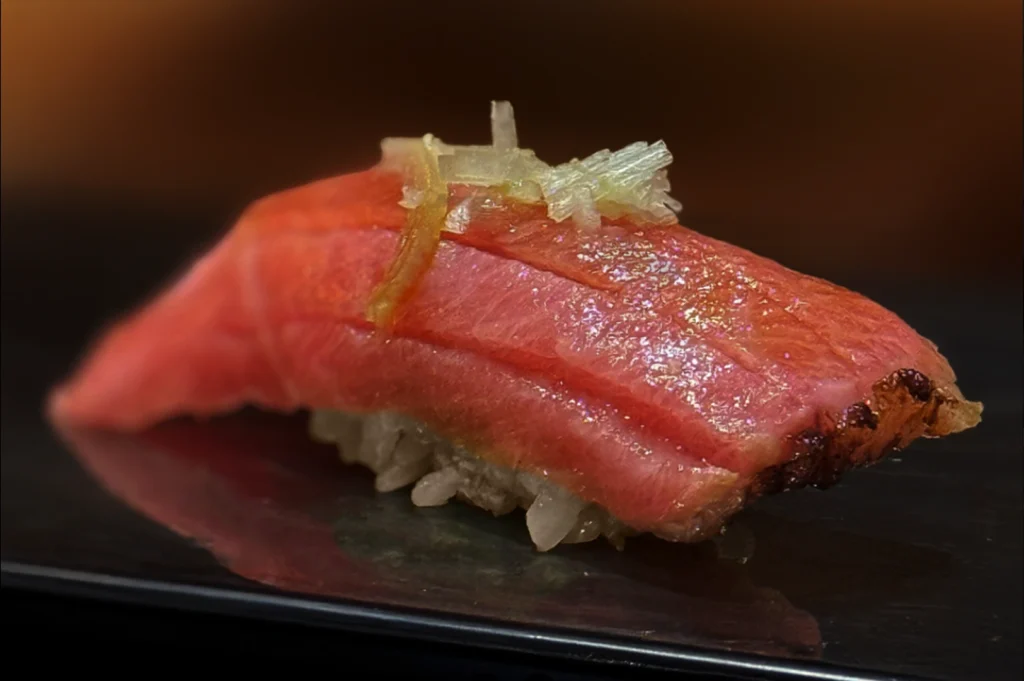 A dramatic, extreme close-up shot features a single piece of premium otoro (fatty tuna) nigiri sushi resting on a reflective black surface against a dark, warm-toned gradient background. The thick, beautifully marbled slice of pink-and-red tuna shows a delicate sear on its edge, a glistening glaze, and a subtle topping of finely shredded ginger or crystal salt flakes, perfectly showcasing its rich texture.
