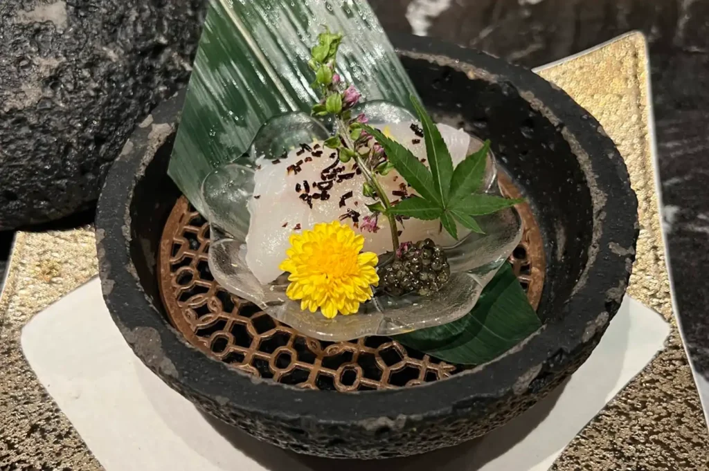 A piece of translucent, pale white sashimi is artfully arranged in a rustic, dark-textured stone bowl. The fish is garnished with a small, vibrant yellow chrysanthemum, a tiny mound of black caviar, and a delicate green maple leaf, all resting on a thin, translucent leaf-shaped plate that sits inside the bowl's patterned metal insert.