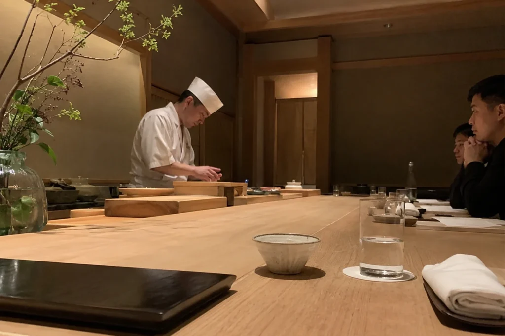 A low-angle shot captures the intimate and serene atmosphere of an upscale omakase sushi bar. On the left, a professional sushi chef wearing a white uniform and traditional tall chef's hat stands behind a pristine, expansive light-wood counter, meticulously preparing food over a wooden cutting board with a delicate potted green branch framing the scene from the far left. On the right, two male customers sit quietly in the dimly lit room, watching the chef with anticipation, while the foreground shows clean, empty ceramic bowls, a glass of water, and a neatly folded white hand towel resting on the smooth wooden surface.