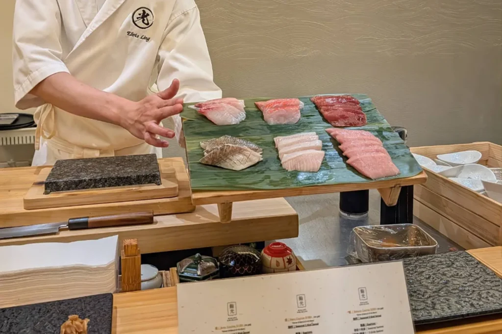 A chef in a crisp white uniform stands behind a wooden counter, gesturing with one hand toward a wooden board holding six precisely sliced pieces of raw fish arranged on a green leaf, showcasing the fresh ingredients prepared for an omakase service.