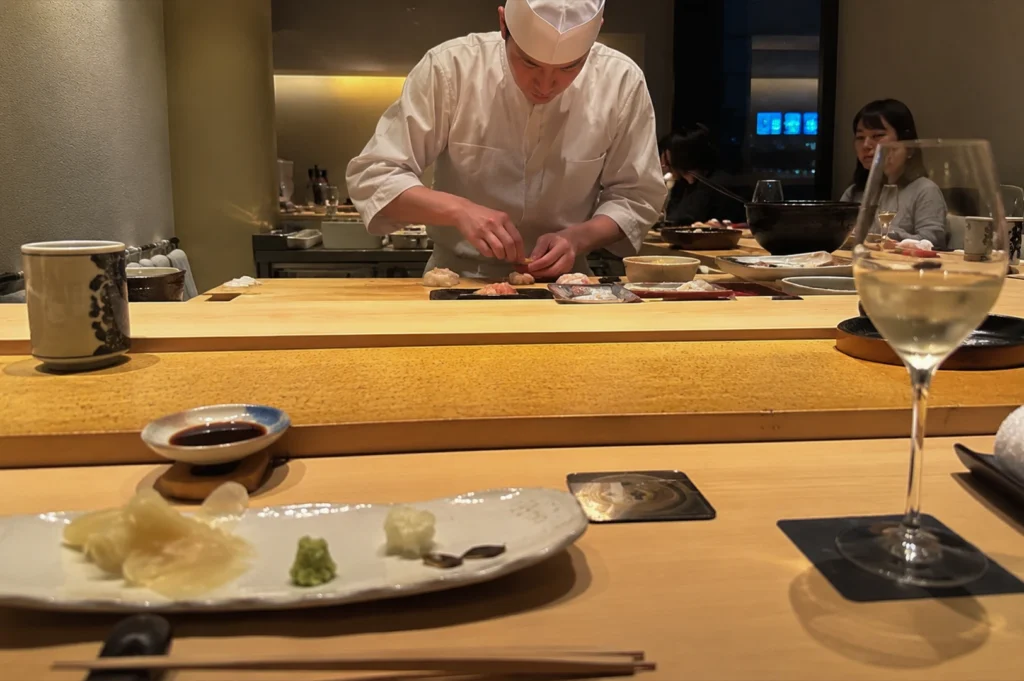 This wide single-panel image shows a chef preparing sushi behind a clean, light-colored wooden omakase counter. The chef, wearing a traditional white uniform and cap, is focused on delicately arranging pieces of nigiri on a serving tray in front of him. In the foreground, a customer's perspective is captured with a long wooden table setting featuring a plate of pickled ginger and wasabi, a small dish of soy sauce, a pair of chopsticks, and a tall glass of white wine catching the ambient light.