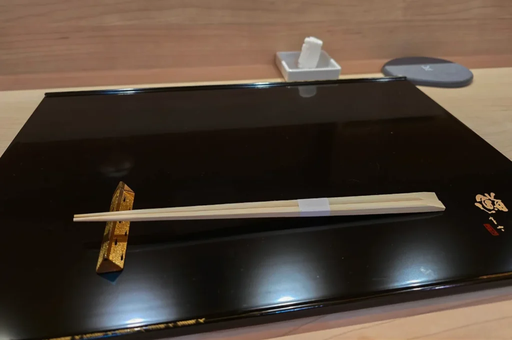 A pristine, minimalist table setting features a glossy black lacquer tray resting on a light-colored wooden counter. On the tray, a pair of light-colored wooden chopsticks is held together with a white paper band and balanced horizontally across a decorative, triangular gold chopstick rest on the left side. A gold-embossed Japanese signature or emblem accents the bottom right corner of the tray, while a small white square dish holding a folded wet towel and a grey circular coaster sit neatly in the background.
