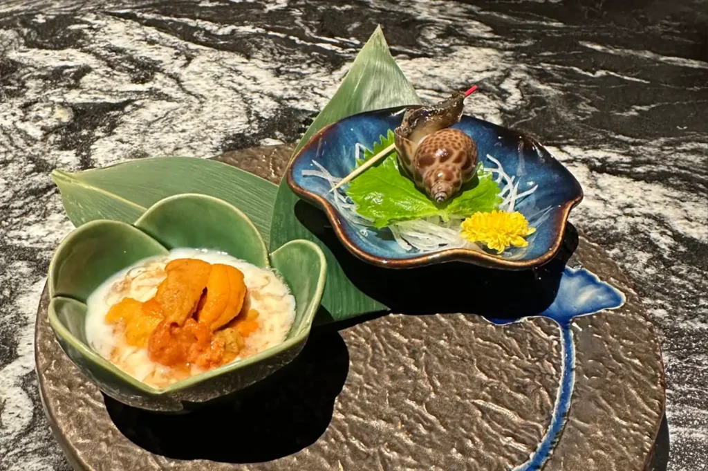 This elevated food presentation features two distinct dishes served on a large, textured dark circular plate: a green, flower-shaped bowl containing creamy uni (sea urchin) nestled in a light sauce, and a deep blue scalloped dish holding a cooked sea snail skewered with a toothpick, accented by a bright yellow chrysanthemum and a bed of daikon radish.