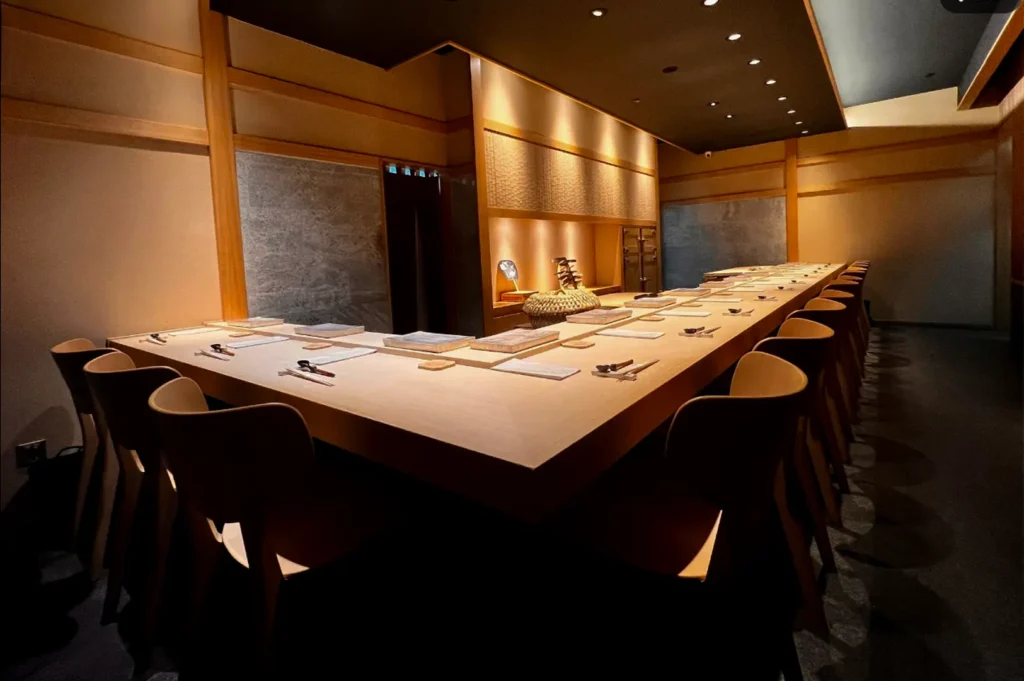 A modern Japanese omakase restaurant featuring a long wooden sushi counter, elegant seating, warm ambient lighting, minimalist décor, and a refined setting for an intimate fine dining experience.