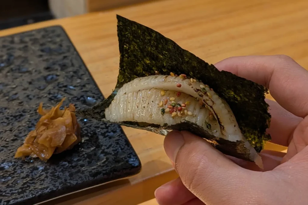A diner holds a hand-rolled temaki sushi piece wrapped in a square of dark nori, revealing a tender, seared slice of seafood topped with colorful sesame seeds, while a small pile of pickled ginger rests on a textured black plate to the left, highlighting the intimate, handcrafted nature of the meal.