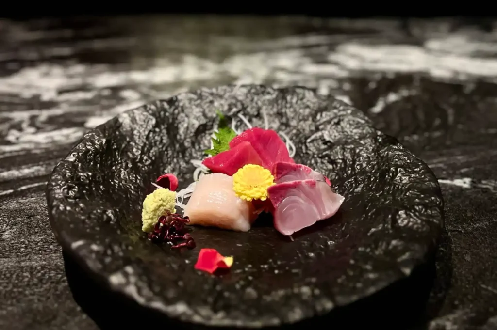 A piece of premium sashimi is elegantly plated in the center of a rustic, unevenly textured matte black dish, featuring vibrant slices of red tuna and paler fish, garnished with a bright yellow chrysanthemum, fresh wasabi, and a few scattered red flower petals against a dark, polished stone countertop background.