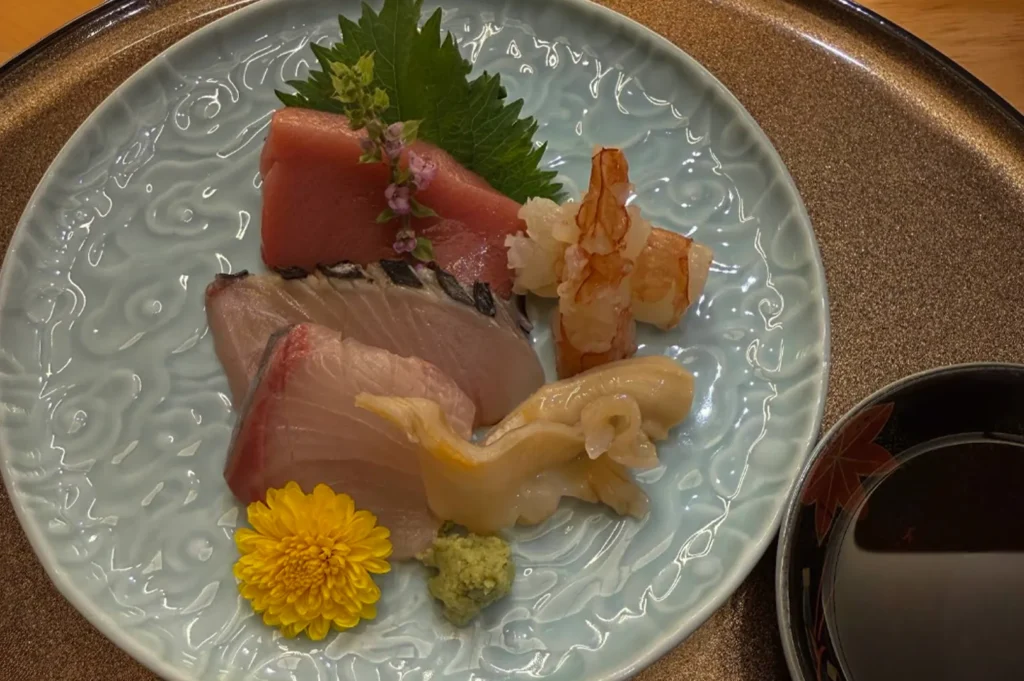 A light blue, textured ceramic plate holds a delicate arrangement of premium sashimi, including thick slices of tuna, a curled prawn, and shellfish, garnished with a bright yellow chrysanthemum, a fresh green shiso leaf, and a small mound of wasabi, presented alongside a dark dipping sauce bowl to emphasize the aesthetic beauty of the course.