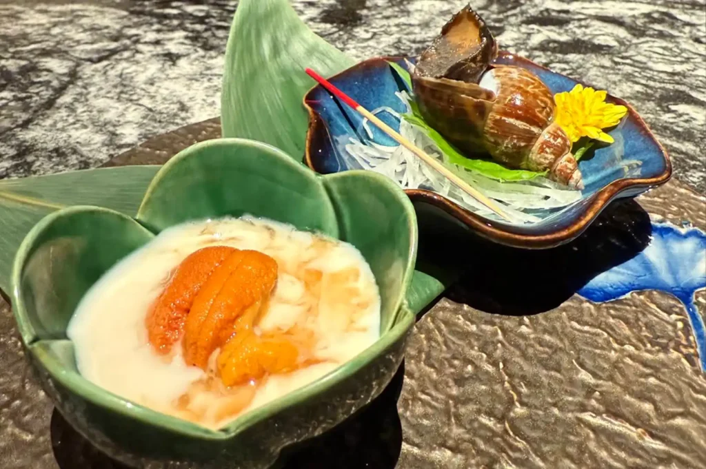 Highlights the contrast between the vibrant orange segments of creamy uni in a soft-toned sauce and the glistening, spiral-shelled sea snail served in a dark blue bowl with a bright yellow flower garnish.