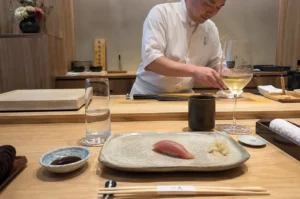 A front-facing, waist-up view behind an omakase bar shows a sushi chef focused on preparing a meal, wearing a crisp white chef's coat behind a wide wooden counter. In the immediate foreground on the guest's side of the counter, a single piece of tuna nigiri sushi sits elegantly on a speckled, rectangular ceramic plate next to a small pile of pickled ginger, flanked by a glass of water, a small dish of soy sauce, a black teacup, a tall wine glass filled with white wine, and a pair of chopsticks wrapped in a paper band resting horizontally.