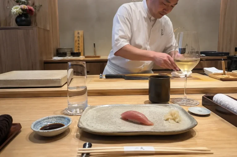 A front-facing, waist-up view behind an omakase bar shows a sushi chef focused on preparing a meal, wearing a crisp white chef's coat behind a wide wooden counter. In the immediate foreground on the guest's side of the counter, a single piece of tuna nigiri sushi sits elegantly on a speckled, rectangular ceramic plate next to a small pile of pickled ginger, flanked by a glass of water, a small dish of soy sauce, a black teacup, a tall wine glass filled with white wine, and a pair of chopsticks wrapped in a paper band resting horizontally.