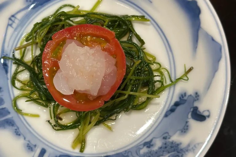 A top-down view shows a slice of a red tomato topped with translucent raw fish placed over a bed of slender green sea asparagus on a white ceramic plate decorated with blue floral patterns.