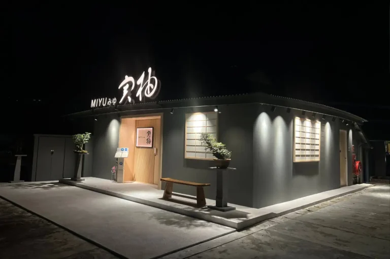 The exterior of the Japanese restaurant MIYU features a sleek dark grey facade softly lit by overhead spotlights at night. A minimalist wooden entrance door is flanked by traditional paper-style shoji window panels, two potted bonsai trees mounted on stands, and a wooden bench resting on the smooth concrete entryway.