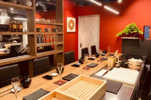 A wide interior shot of a minimalist, elegant sushi restaurant featuring a long wooden counter with black chairs. The walls are a striking, deep red, accented by a wooden shelf holding glassware and bottles, a small bonsai tree on a shelf, and clean, modern decor that creates an inviting, upscale atmosphere.