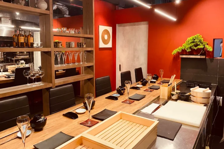 A wide interior shot of a minimalist, elegant sushi restaurant featuring a long wooden counter with black chairs. The walls are a striking, deep red, accented by a wooden shelf holding glassware and bottles, a small bonsai tree on a shelf, and clean, modern decor that creates an inviting, upscale atmosphere.