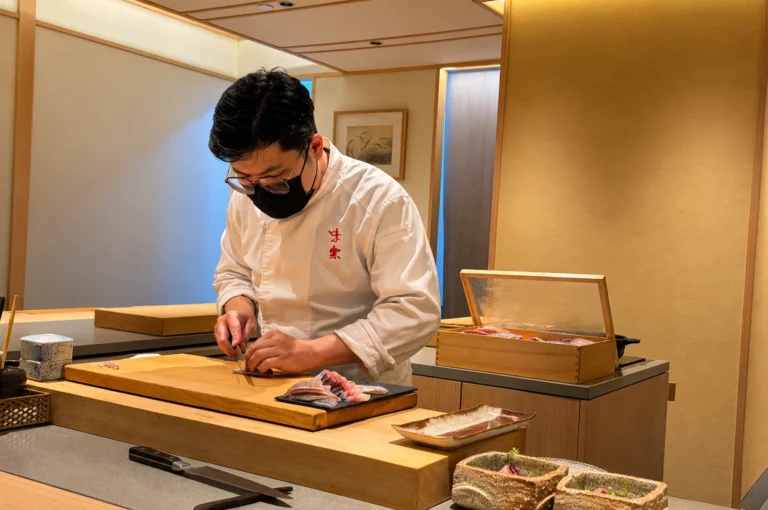 A professional chef wearing a white uniform and black face mask stands behind a long, wooden counter, meticulously slicing pieces of raw fish on a wooden board at a high-end Japanese restaurant. He is surrounded by various kitchen tools and containers, and the minimalist, warmly lit interior features wood paneling that creates a serene and focused culinary environment.