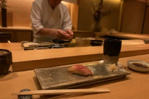 In an intimate, warmly lit Japanese sushi restaurant, a setting for an omakase meal is arranged on a light wood counter. In the foreground, a single piece of fresh nigiri sushi featuring a glistening slice of pink fish rests on a rectangular metallic ceramic platter alongside a small pile of pickled ginger. A pair of wooden chopsticks rests on a patterned stone holder in front, accompanied by a small dish of soy sauce, a dark ceramic teacup, and a lid container. In the softly blurred background, a sushi chef wearing a white chef's coat stands behind the raised prep area, meticulously hand-shaping sushi.