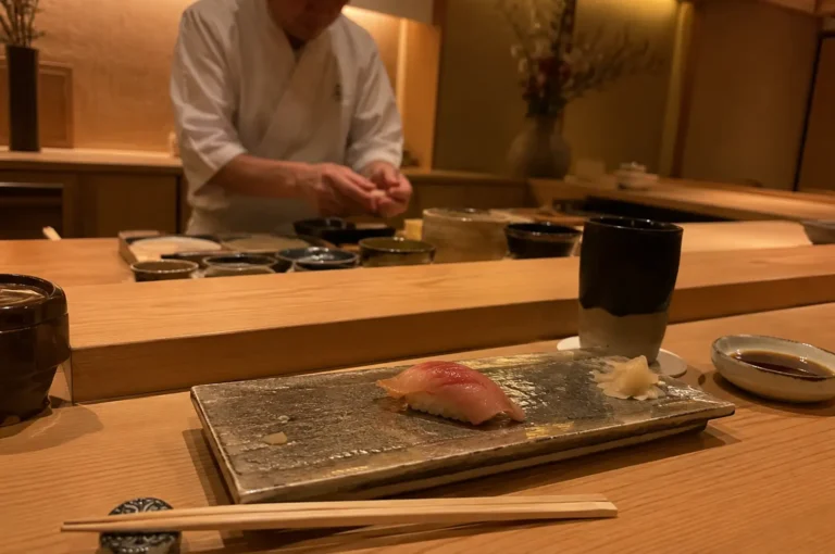 In an intimate, warmly lit Japanese sushi restaurant, a setting for an omakase meal is arranged on a light wood counter. In the foreground, a single piece of fresh nigiri sushi featuring a glistening slice of pink fish rests on a rectangular metallic ceramic platter alongside a small pile of pickled ginger. A pair of wooden chopsticks rests on a patterned stone holder in front, accompanied by a small dish of soy sauce, a dark ceramic teacup, and a lid container. In the softly blurred background, a sushi chef wearing a white chef's coat stands behind the raised prep area, meticulously hand-shaping sushi.