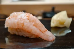 A single piece of nigiri sushi, featuring a pale, translucent shrimp draped over vinegared rice, sits on a dark, glossy serving board next to a small mound of pickled ginger.