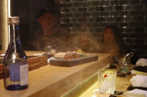 An atmospheric, eye-level shot captures a dining scene at a wooden omakase counter where a fine, golden mist or seasoning dust floats through the air under a warm spotlight. In the center, an artisanal dark stone slab displays a beautifully arranged gourmet dish. In the foreground, a tall, clear glass bottle of sake with a blue label sits on the left, balanced by a textured crystal glass with a slice of lemon on the right. In the softly blurred background, two diners sit behind the counter against a dark, tiled wall, watching the chef's presentation.
