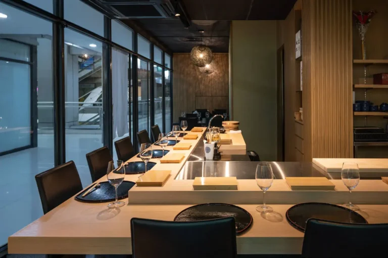 A sleek, modern sushi restaurant features a long, L-shaped light wood dining counter lined with minimalist black leather chairs. Each seating arrangement is neatly set with a textured black circular plate, a wooden block serving stand, and an empty wine glass under the warm glow of spherical mesh pendant lights. The minimalist space features light wood-slatted walls, while a floor-to-ceiling glass wall on the left looks out onto a brightly lit indoor commercial corridor or mall interior.