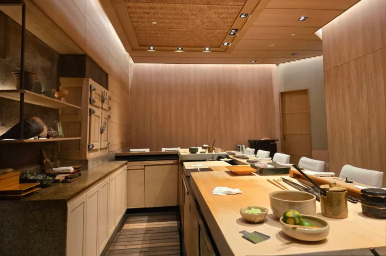 An intimate, upscale Japanese omakase restaurant interior featuring warm, light-toned wooden walls, a woven ceiling panel, and a long wooden chef’s preparation counter arranged with ceramic bowls, fresh citrus, and prep tools, facing a row of high-backed dining chairs.