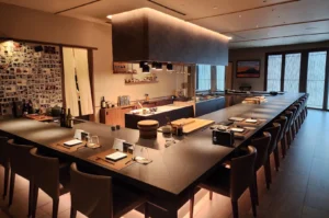 A wide, inviting view of a modern, upscale Japanese Kappou restaurant's interior featuring a large, dark U-shaped wooden counter that wraps around a central open kitchen. Warm, ambient light glows from under the counter and from a large, rectangular hood hanging above the preparation area where wooden cutting boards, containers, and chef tools are neatly arranged. Elegant high-backed leather chairs line the counter, which is set with individual placemats, menus, glassware, and a black luxury handbag. The background showcases sliding wooden slatted doors and a wall collage filled with polaroid photos of smiling guests.