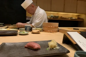 The image captures a serene, high-end sushi dining experience featuring a skilled chef in a white coat and traditional hat, focused intently on preparing dishes behind a long, light-wood counter. In the foreground, a single, beautifully prepared piece of pink tuna nigiri is served alongside a small mound of pickled ginger on a textured, dark rectangular ceramic plate. The counter is elegantly minimalist, adorned with various small, traditional ceramic dishes, a lidded wooden container for rice, and subtle culinary tools, all set against a backdrop of warm, ambient lighting and wooden cabinetry that enhances the sophisticated, quiet atmosphere of an authentic omakase restaurant.