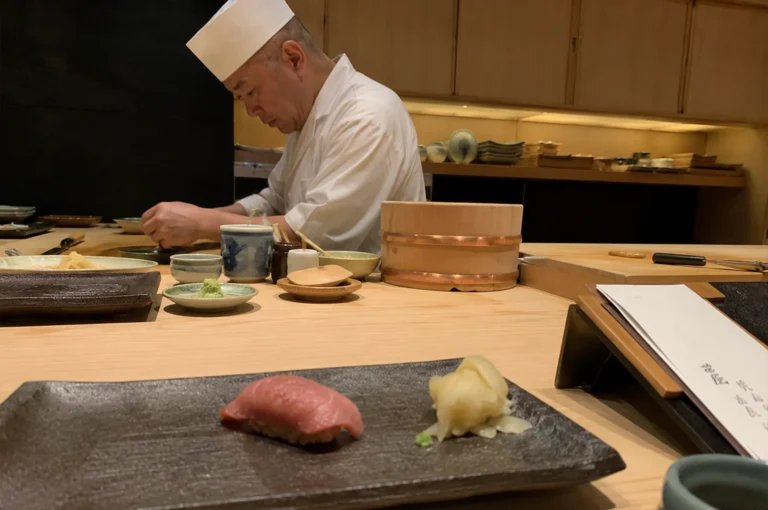 The image captures a serene, high-end sushi dining experience featuring a skilled chef in a white coat and traditional hat, focused intently on preparing dishes behind a long, light-wood counter. In the foreground, a single, beautifully prepared piece of pink tuna nigiri is served alongside a small mound of pickled ginger on a textured, dark rectangular ceramic plate. The counter is elegantly minimalist, adorned with various small, traditional ceramic dishes, a lidded wooden container for rice, and subtle culinary tools, all set against a backdrop of warm, ambient lighting and wooden cabinetry that enhances the sophisticated, quiet atmosphere of an authentic omakase restaurant.