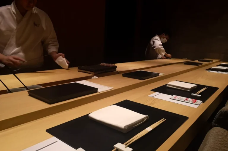 A sushi chef in a white uniform and face mask works at a long wooden counter, preparing food while another person works in the background, with black rectangular plates and folded white napkins neatly arranged for diners.