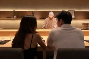 An intimate dining experience unfolds from a reverse angle inside a high-end, minimalist Japanese omakase sushi restaurant. In the foreground, a couple sits with their backs to the camera at a light-colored, pristine wooden counter; on the left, a woman with long, flowing dark hair wears a black spaghetti-strap dress, while on the right, a man with short dark hair wears a crisp white short-sleeved polo shirt. Across the wide wooden counter in the center background, a professional sushi chef clad in a traditional white jacket and matching chef's hat stands under soft, ambient cove lighting, looking downward with deep concentration as he meticulously prepares food. The sleek background features a recessed wooden display shelf adorned with minimalist decor, including a small floral arrangement in a white vase, a sake bottle, and a framed art piece, all set beneath a warm, textured wall and a ceiling lined with elegant wooden slats.