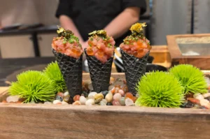A close-up shot highlights three gourmet temaki hand rolls styled like miniature ice cream cones, standing upright in a bed of smooth, multi-colored river pebbles inside a rustic wooden box. The cones are made from dark, checkered seaweed sheets and are filled to the brim with a rich, pink tuna tartare mixed with orange salmon roe, then garnished with chopped chives, black caviar, and a delicate topping of shimmering gold leaf. Sprigs of vibrant green, faux moss-like plants flank the cones on either side, while a blurred chef stands in the background.