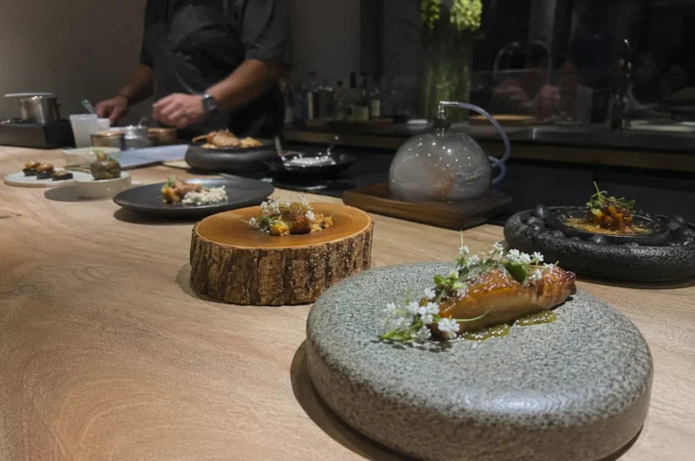 A dimly lit, high-end fine dining counter made of smooth light wood displays an array of artfully plated gourmet dishes stretching into the background. In the immediate foreground, a glazed fillet of fish decorated with delicate white flowers sits on a round, thick grey stone plate. Next to it, a small morsel rests atop a natural, rustic tree stump slab. Further down the counter, other plated courses are arranged on stone, ceramic, and wooden dishware—including one covered with a glass cloche filled with trapped smoke—while a chef in a dark uniform works out of focus in the background.