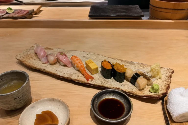 An eye-level, unfiltered lifestyle shot captures a premium selection of sushi and side dishes neatly arranged on a light-colored wooden dining counter. The main assortment rests on an elongated, rustic ceramic platter with wavy edges and features various types of nigiri, including pale pink fish, striped ebi (shrimp), a block of yellow tamago (egg omelet), two pieces of gunkan maki topped with orange roe, scallop nigiri wrapped in nori, and a piece of grilled white fish. A small mound of pickled ginger and a dollop of green wasabi sit at the right end of the platter. In the foreground, several small matching ceramic dishes hold hot green tea, pickled slices, and dark soy sauce, while a rolled white hand towel rests on a small tray to the right.