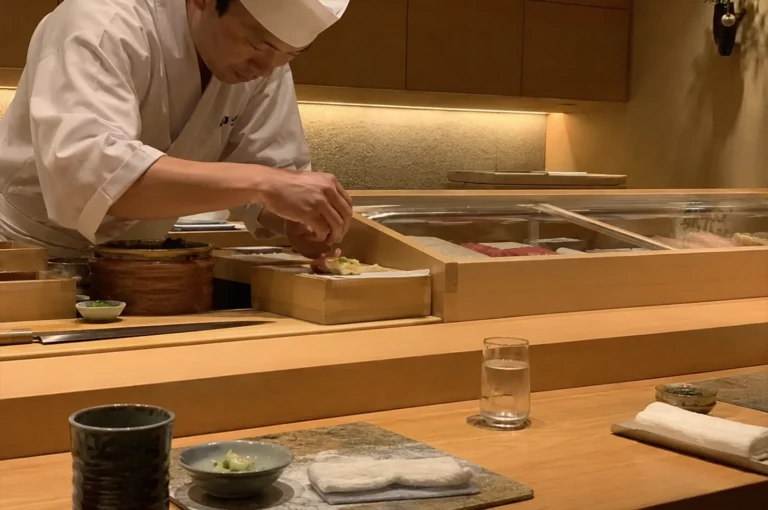 A close-up perspective from a diner's seat captures a sushi chef meticulously crafting a dish behind a pristine, light-colored wooden omakase counter. In the foreground, a customer's table setting features a textured dark ceramic tea cup, a small grey dish containing grated wasabi, and a stone serving plate with a folded white towel, while a clear glass of water rests further to the right. In the midground, the chef—clad in a traditional white jacket and matching chef's hat—leans forward with intense focus, using both hands to delicately assemble a piece of sushi on a raised wooden platform. A long, slender yanagiba sushi knife lies horizontally on the counter to his left, and a long, glass-topped wooden display case showcasing fresh blocks of fish stretches behind his work area. The background radiates a warm, elegant minimalism, featuring light wood wall panels, a textured backsplash highlighted by soft cove lighting, and a subtle hanging vase with fresh green foliage on the far right.