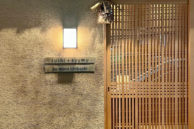 The entrance to Sushi Ayumu features a sophisticated and minimalist design, with a textured, warm-toned stone wall and a sliding door crafted from vertical light-wood slats. A soft, glowing rectangular wall sconce illuminates the signage, which clearly displays "sushi · ayumu" and "by masa ishibashi," while a small, traditional Japanese straw decoration hangs above the entrance, signaling an authentic dining experience.