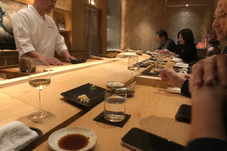 The image features the warm, sophisticated interior of an omakase restaurant, where a smiling chef in a white uniform stands behind a long, pristine light-wood counter. Several patrons are seated along the counter, engaged in conversation while enjoying their meal, with small plates of food, glasses of wine, and water placed before them. The scene is illuminated by soft, ambient lighting that reflects off the wooden surfaces and minimalist decor, creating an intimate and exclusive atmosphere. In the immediate foreground, a plate of soy sauce, a white towel, and a smartphone are laid out on the table, emphasizing the personal and focused nature of the high-end dining experience.