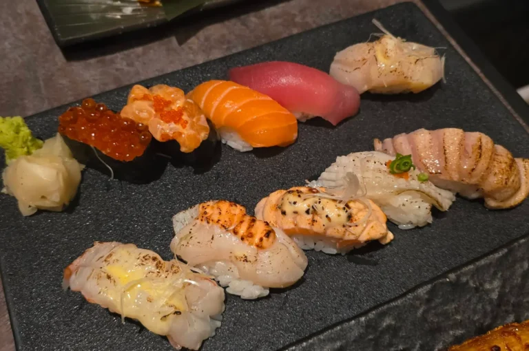 A long black slate stone displays an array of diverse nigiri sushi, including seared scallop, salmon with roe, and various white fish topped with delicate garnishes like scallions, arranged neatly alongside a small pile of pickled ginger and freshly grated wasabi.