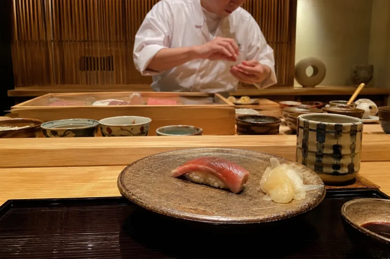 A close-up perspective from a Japanese sushi bar dining setting, showcasing a single piece of tuna nigiri sushi and a small cluster of pale pickled ginger (gari) presented on a textured, circular brown ceramic plate. To the right of the plate sits a tall, cylindrical ceramic tea cup with a hand-painted blue and black checkered grid pattern, all resting on a dark, glossy lacquered tray. In the softly focused background, a sushi chef in a clean white uniform carefully shapes sushi behind a light wooden counter lined with several ceramic bowls, ingredient boxes, and a vertical wooden slat wall.