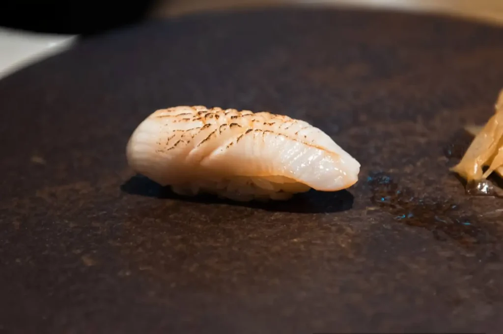 A single piece of aburi nigiri sushi with lightly blowtorched white fish rests on a dark, textured stone platter next to a portion of pickled ginger.