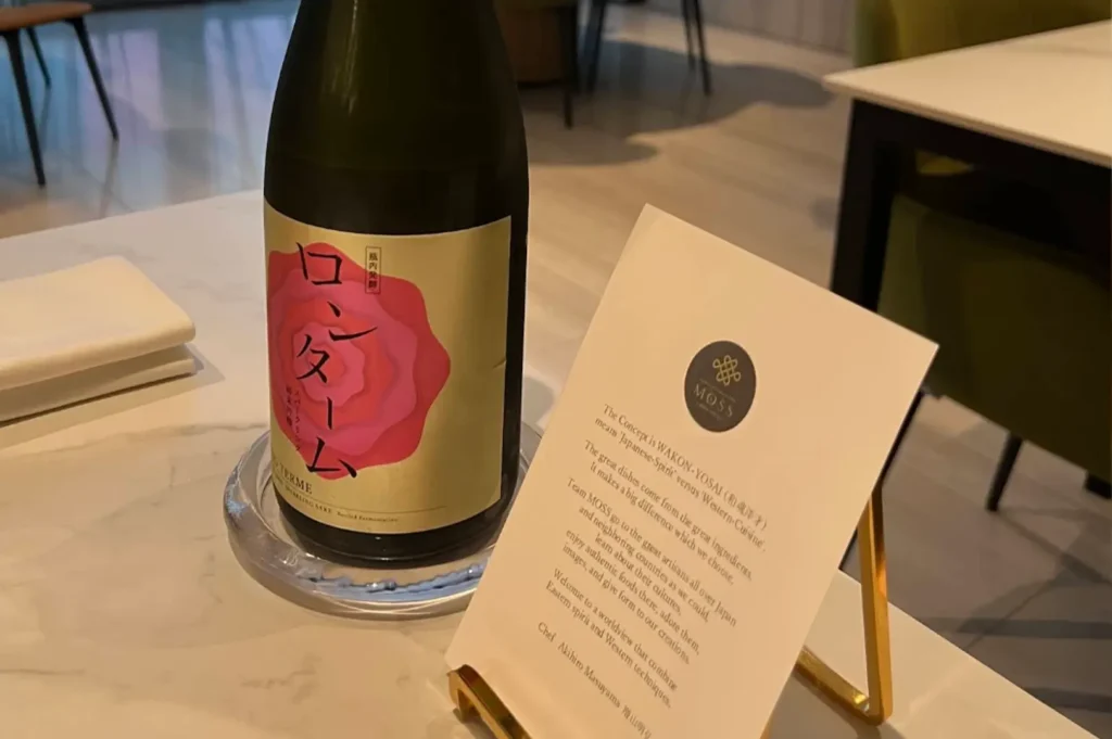 A bottle of sparkling sake with a prominent pink floral label sits on a white marble surface next to a small, framed card that introduces the restaurant's "Wakon-Yosai" concept, which fuses Japanese spirit with Western cuisine.
