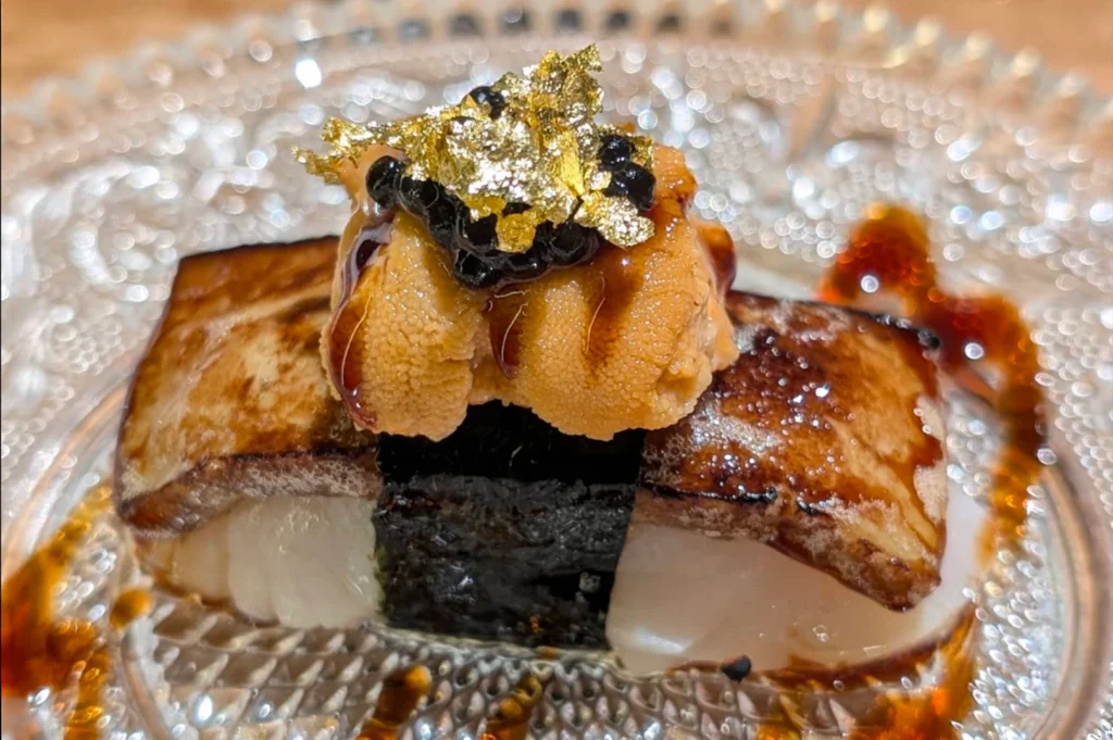 A piece of nigiri sushi, featuring a seared topping and a dollop of sea urchin garnished with black caviar and edible gold leaf, rests on a decorative, clear glass plate.