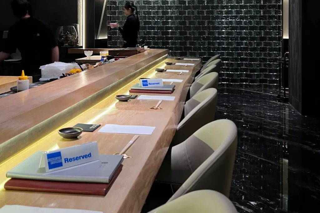 A sleek, modern sushi bar features a long light-wood dining counter lined with stylish beige armchairs, under-counter ambient lighting, dark marble flooring, and "Reserved" signs placed at individual settings.