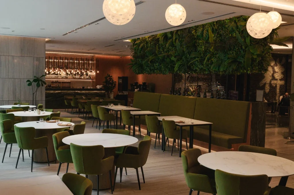 The spacious, elegant interior of a restaurant features circular white marble tables surrounded by upholstered olive-green chairs, with a massive, lush ceiling installation of hanging ferns and glowing, orb-shaped pendant lights overhead.