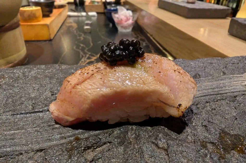 A piece of seared fatty tuna nigiri crowned with a mound of glossy black caviar sits on a dark grey slate serving board at a sushi counter.