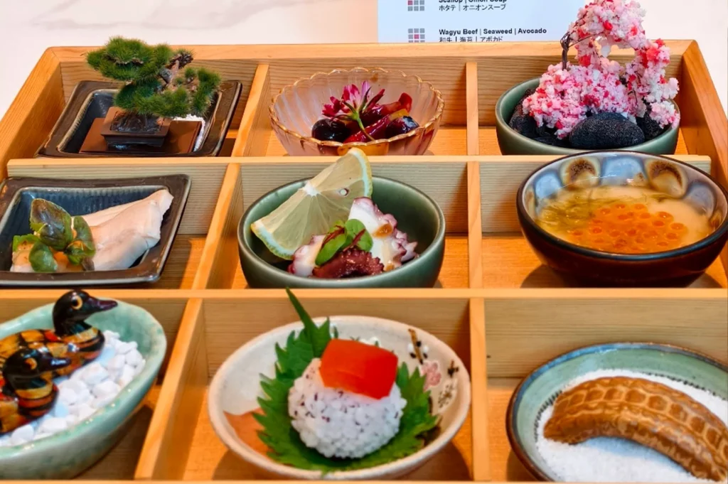 A wooden tray is partitioned into several sections, each containing a small, elegant appetizer, including a tiny artificial bonsai tree, a bowl of octopus with lemon, a glass dish of roe, and a rice ball topped with red garnish, set against a blurred background.