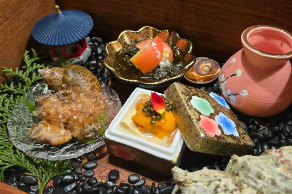 An elaborate assortment of appetizers arranged inside a wooden display box layered with smooth black river pebbles and green pine needles. The presentation includes a small glass dish with sea snail meat topped with dancing bonito flakes near its shell, a scalloped gold bowl holding dark seaweed (mozuku) topped with tomato wedges, a square patterned box containing a creamy mixture topped with vibrant orange sea urchin and caviar, a miniature pink ceramic sake jug, and a tiny decorative Japanese pagoda model on the left.